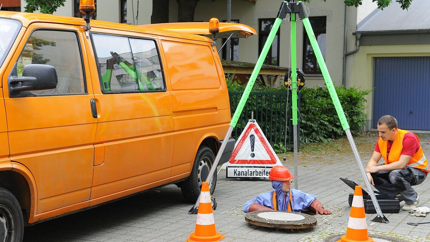Blick auf geöffneten Kanaldeckel, darüber aufgebauten Dreibock und gesicherten Mitarbeiter, der in den Schacht einsteigt